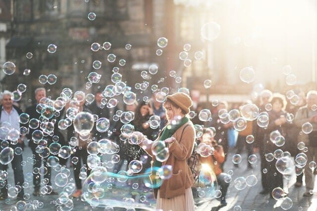 Outdoor scene, woman in fuzzy brown had and sweater with a green scarf playing with many bubbles in front of a crowd of adults